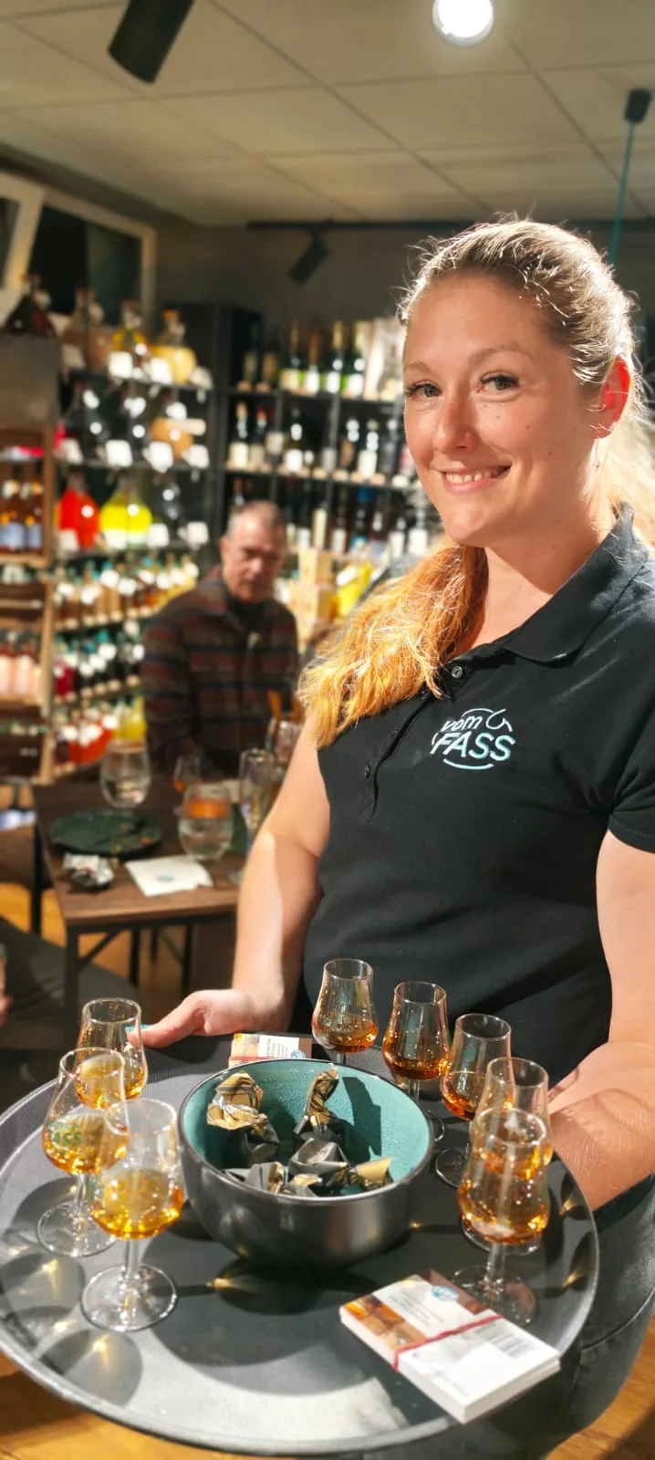 vomFASS-Mitarbeiterin serviert ein Tablett mit Nosing-Gläsern voll Whisky vor der Flaschenwand im Laden